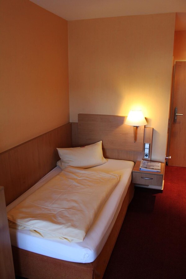 A hotel room with a single bed, a nightstand with a lamp, and a door. - Hotel an der Ilse (Lemgo)