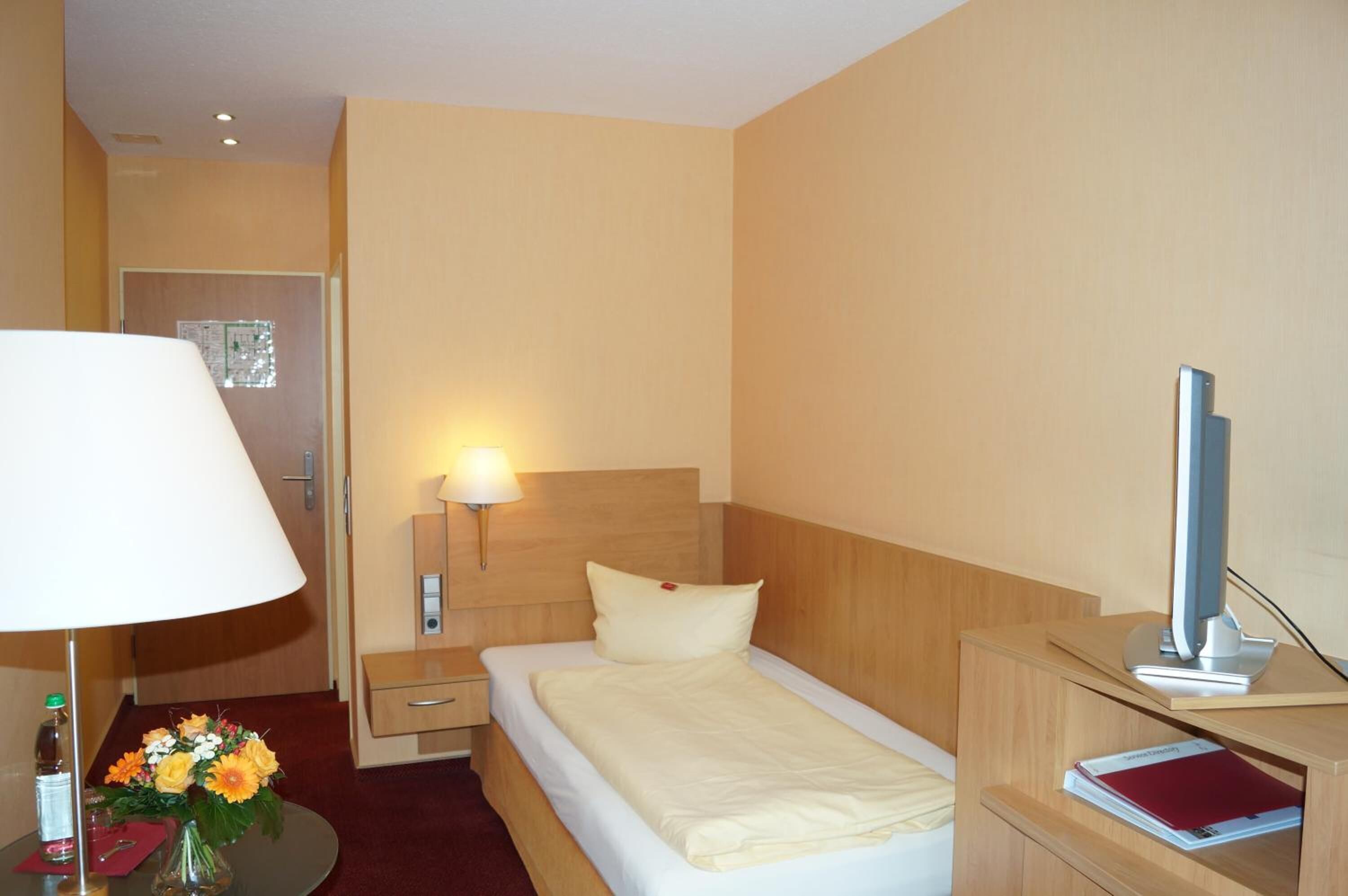 A hotel room with a bed, bedside table, lamp, and a desk with a computer.