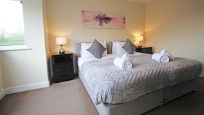 A neatly made bed with a grey patterned bedspread and pillows, two bedside lamps, a wall-mounted abstract painting, and a window with a view of greenery. - 3 Bedroom Holiday Home With River Views (Frodsham)