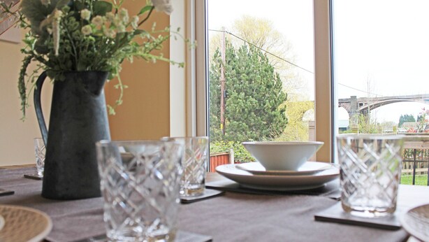 A dining table with a vase of flowers, clear glassware, and plates, set against a window with a view of a bridge and greenery. - 3 Bedroom Holiday Home With River Views (Frodsham)