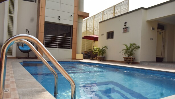 A swimming pool with a metal ladder, a blue chair, and a red umbrella. - Qualibest Grand Hotels (Abuja)