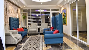 A modern living room with a large blue sofa, grey armchairs, and a patterned rug. - Qualibest Grand Hotels (Abuja)