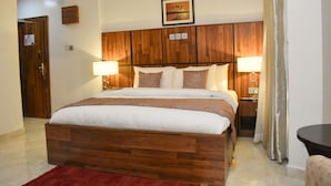 A hotel room with a wooden headboard, a large bed with white bedding, two bedside lamps, and a small table with a chair. - Qualibest Grand Hotels (Abuja)