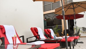 A row of lounge chairs with red cushions and white towels, a glass table with a drink, and a large umbrella. - Qualibest Grand Hotels (Abuja)