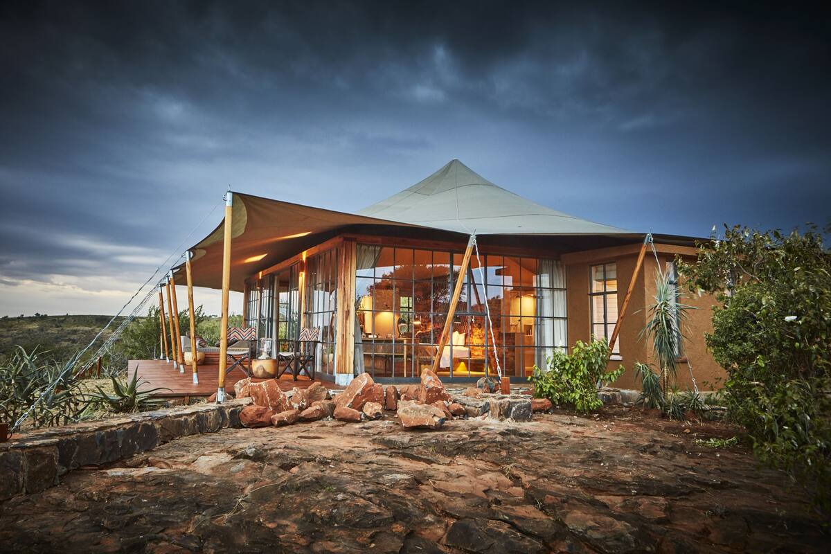 Un lodge moderno y espacioso con un amplio muro de cristal, entrada empedrada y paisaje natural circundante.