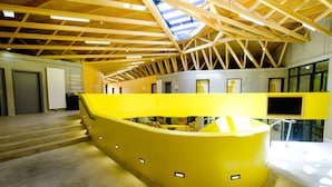 Un interior moderno con un mostrador de recepción amarillo curvo, vigas de techo de madera y una escalera que conduce a niveles superiores.