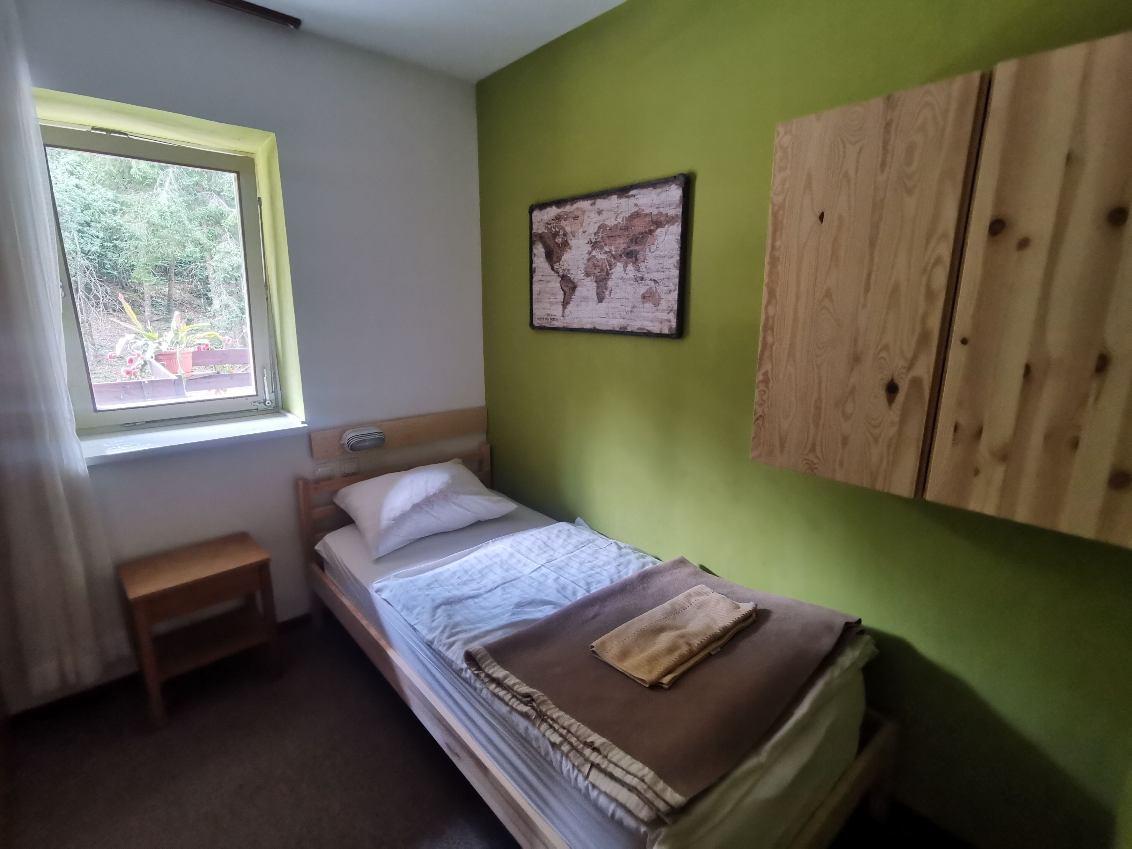 Habitación pequeña con cama individual, ventana, mesita de noche, un mapa en la pared y un armario de madera.