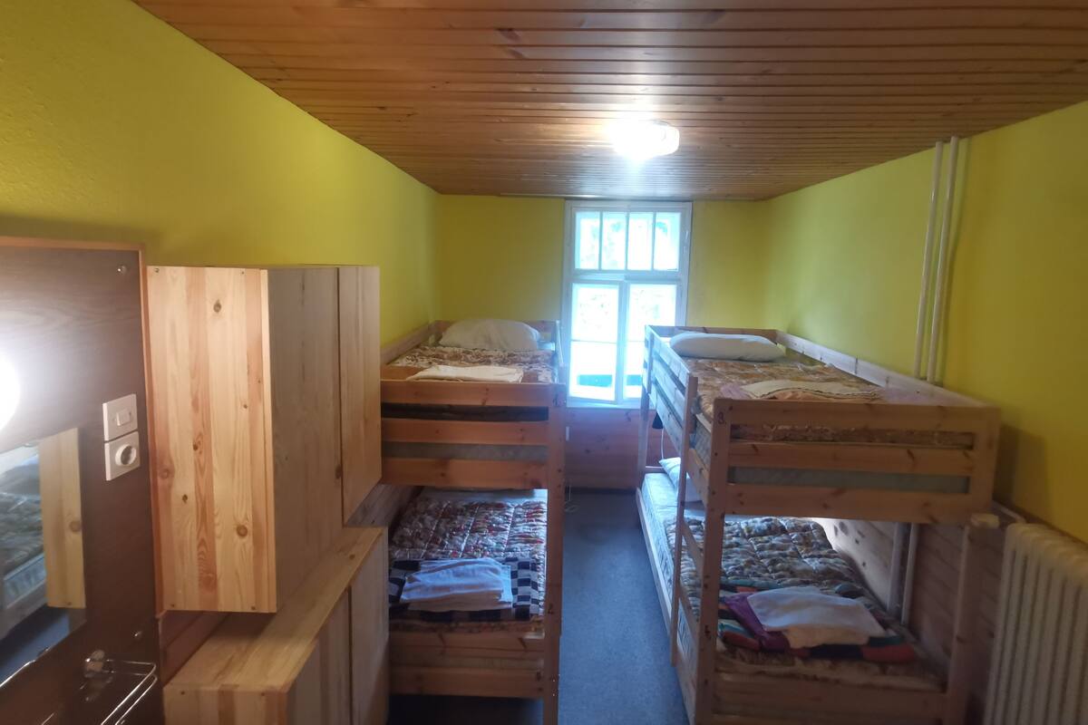 A dormitory room with bunk beds, a wooden wardrobe, and a window with natural light.