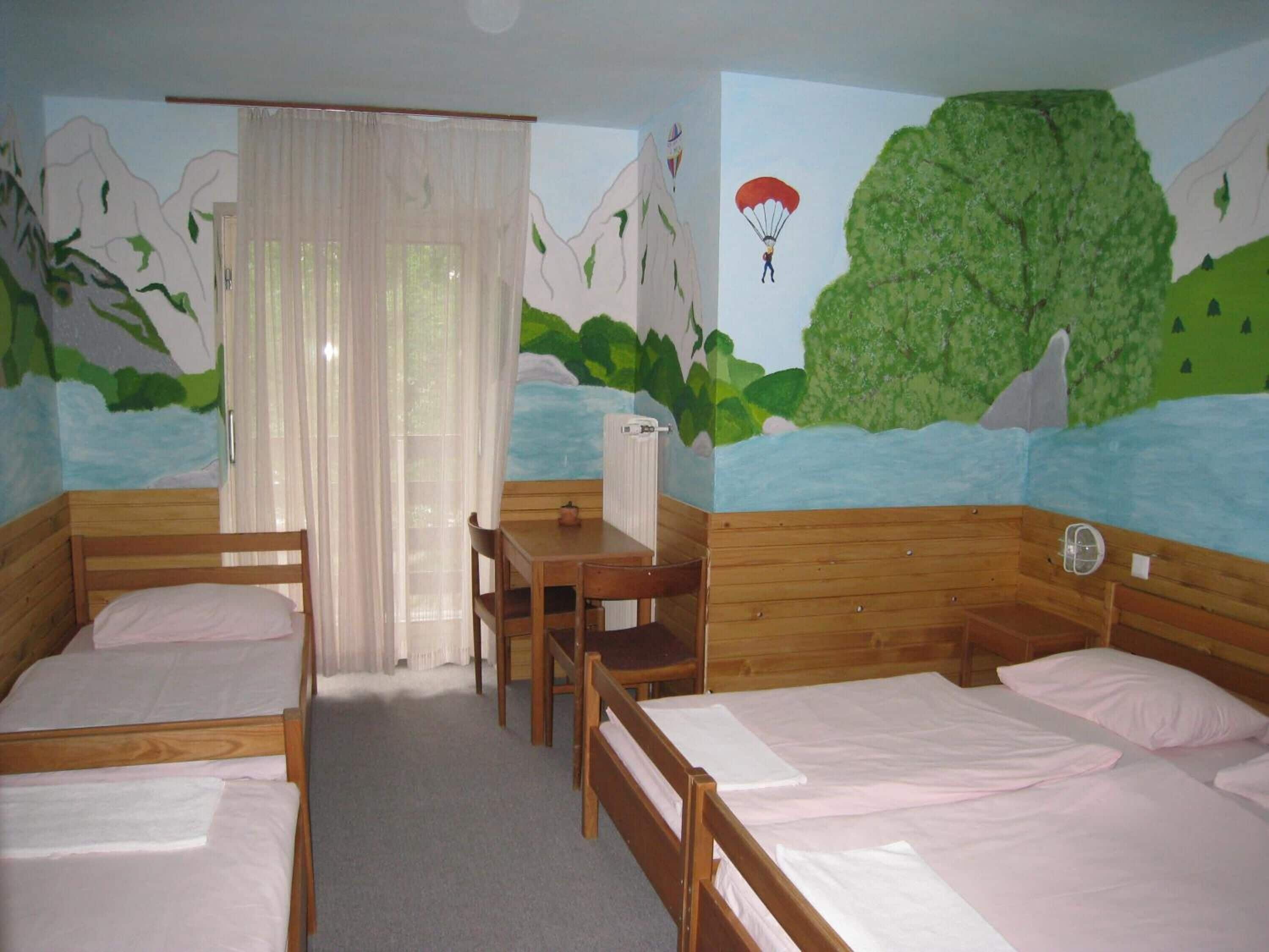 Een kamer met twee houten bedden, een houten bureau en een stoel. Er is een raam met dunne gordijnen, een radiator en een muurschildering van een landschap met bergen en een parachutist.