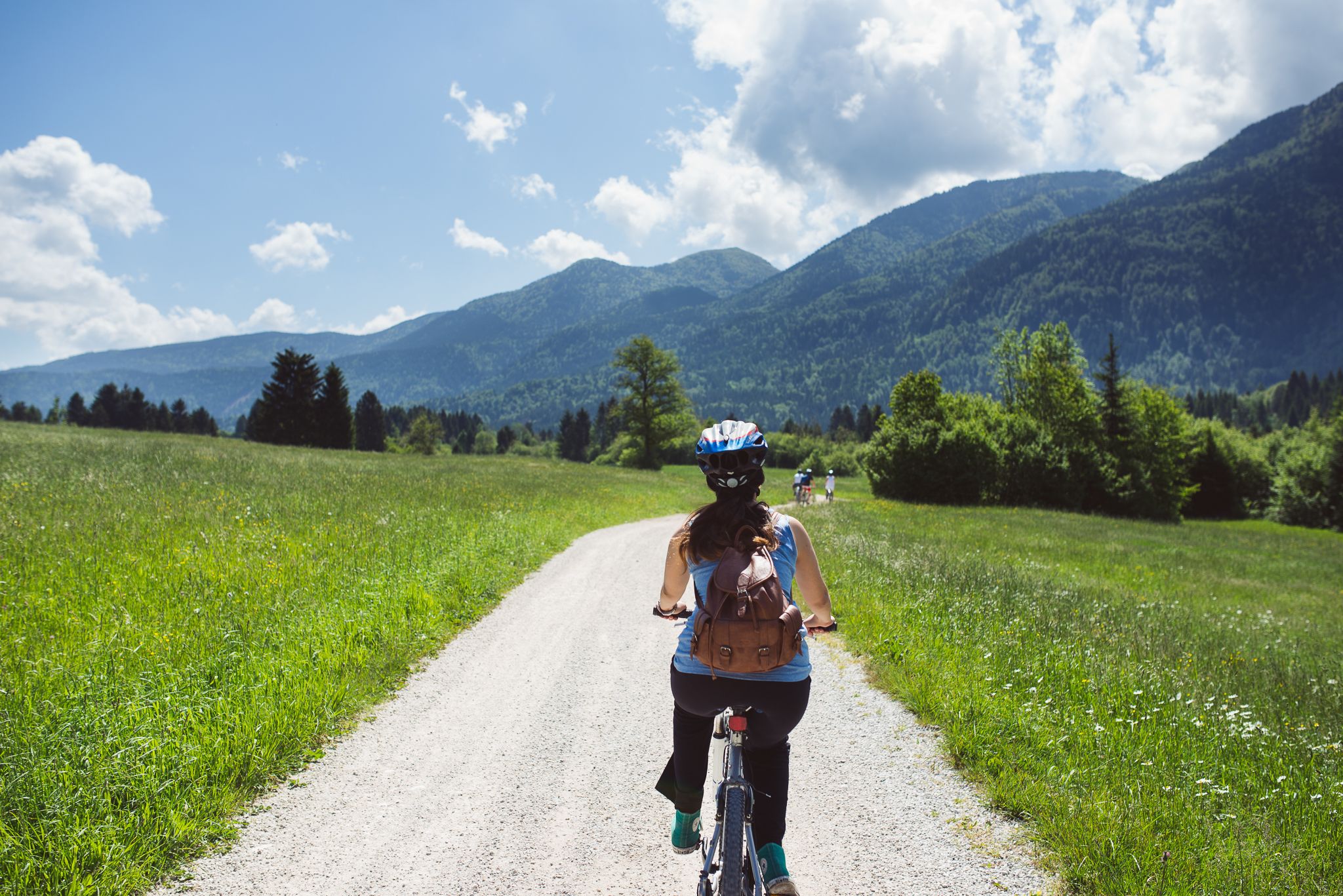 A person riding a bicycle on a gravel path surrounded by green fields and mountains.