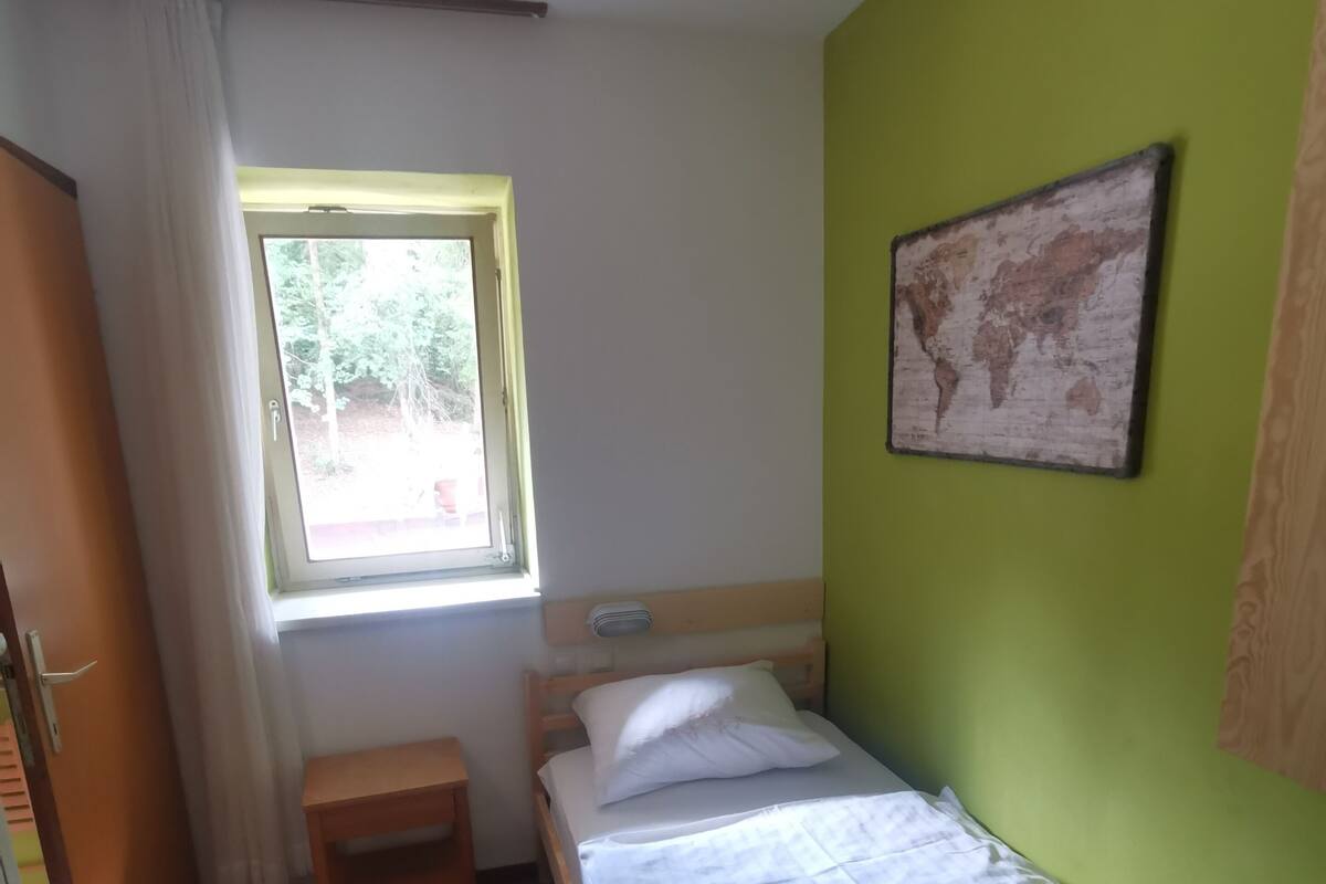A small, single-bed room with a window, a bedside table, a map on the wall, and a door to the left.