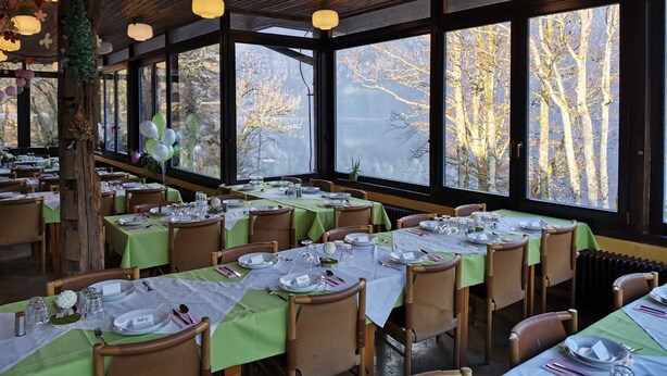 A restaurant with multiple tables set for dining, large windows offering views of trees and a lake, and wooden ceiling beams.