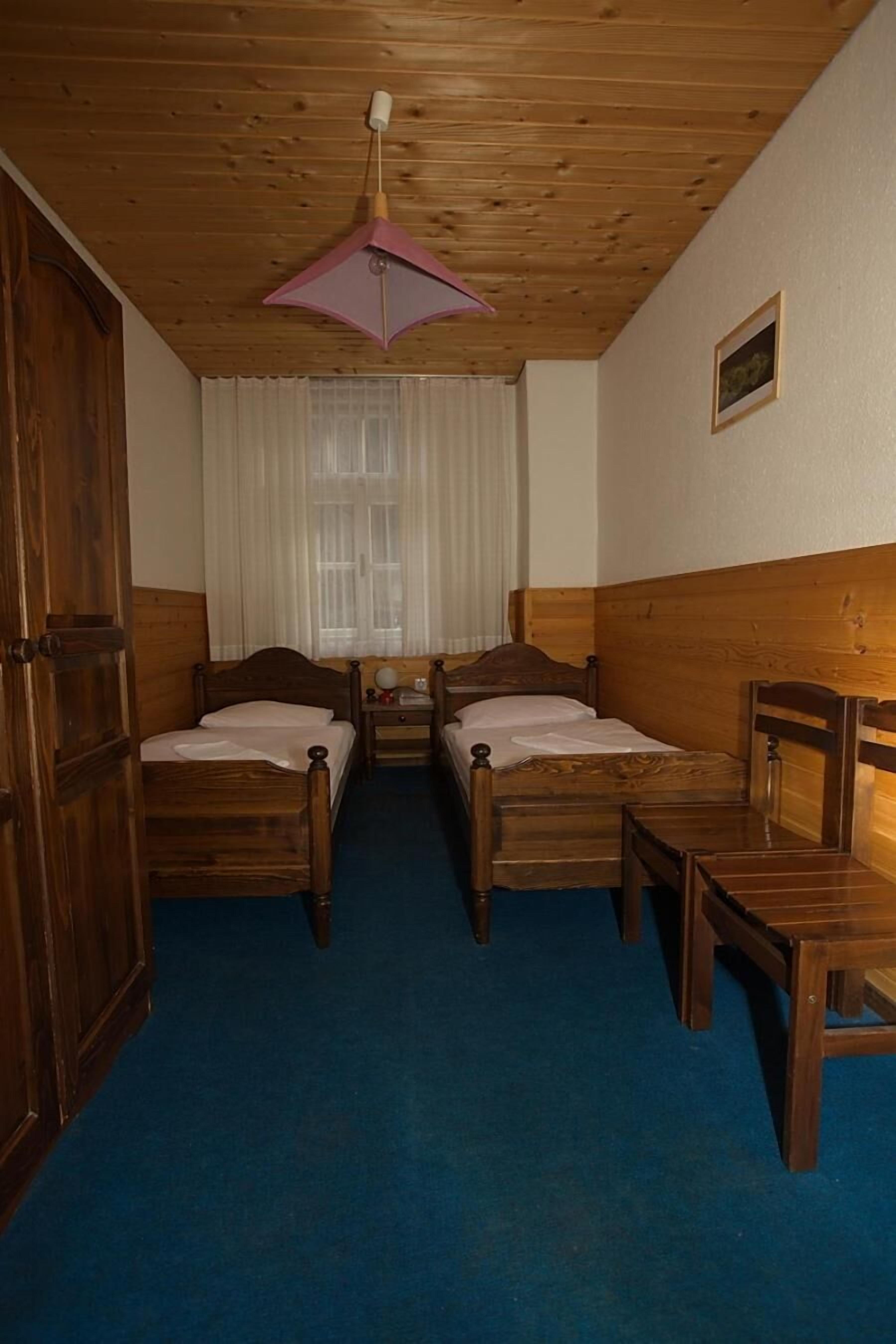 A room with two wooden beds, a wooden chair, a small table, a wooden wardrobe, and a ceiling fan.
