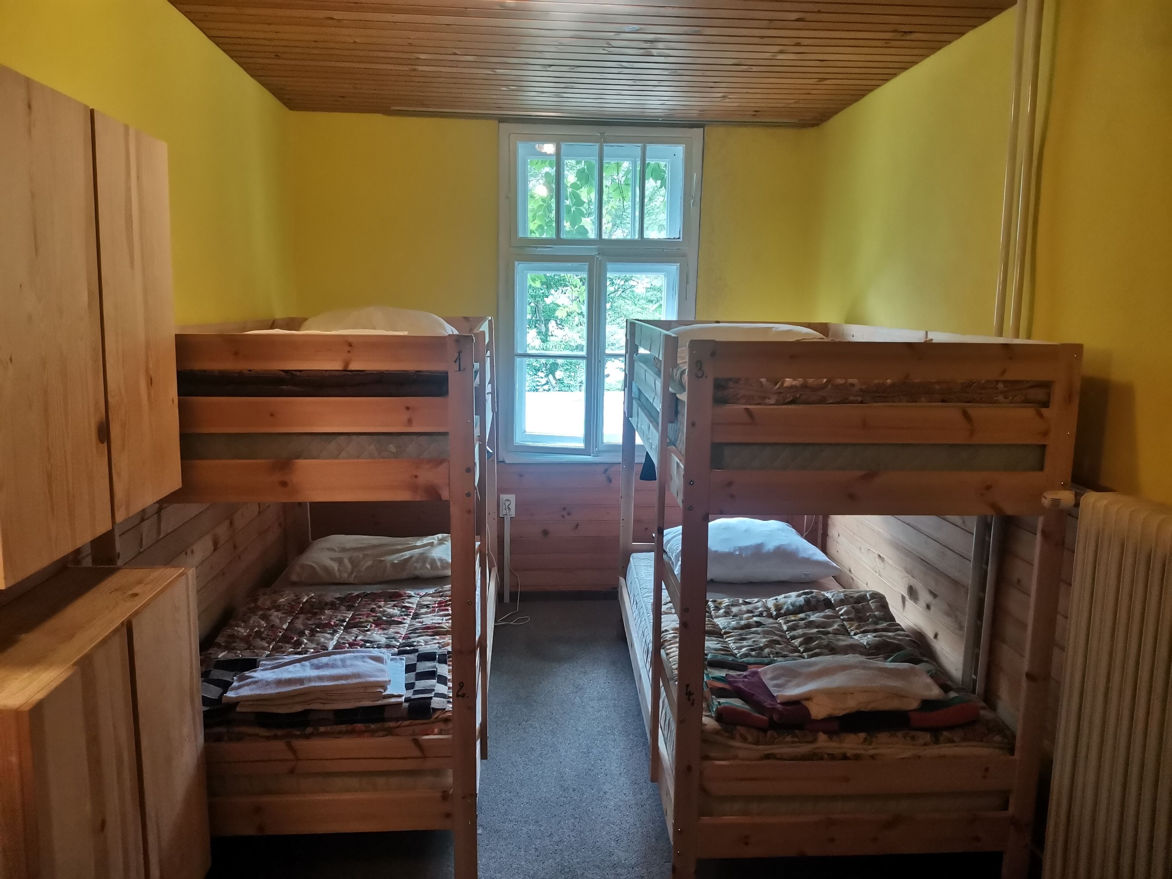 Une chambre de dortoir avec des lits superposés, des murs en bois, une fenêtre donnant sur un paysage verdoyant et un radiateur sur le côté droit.