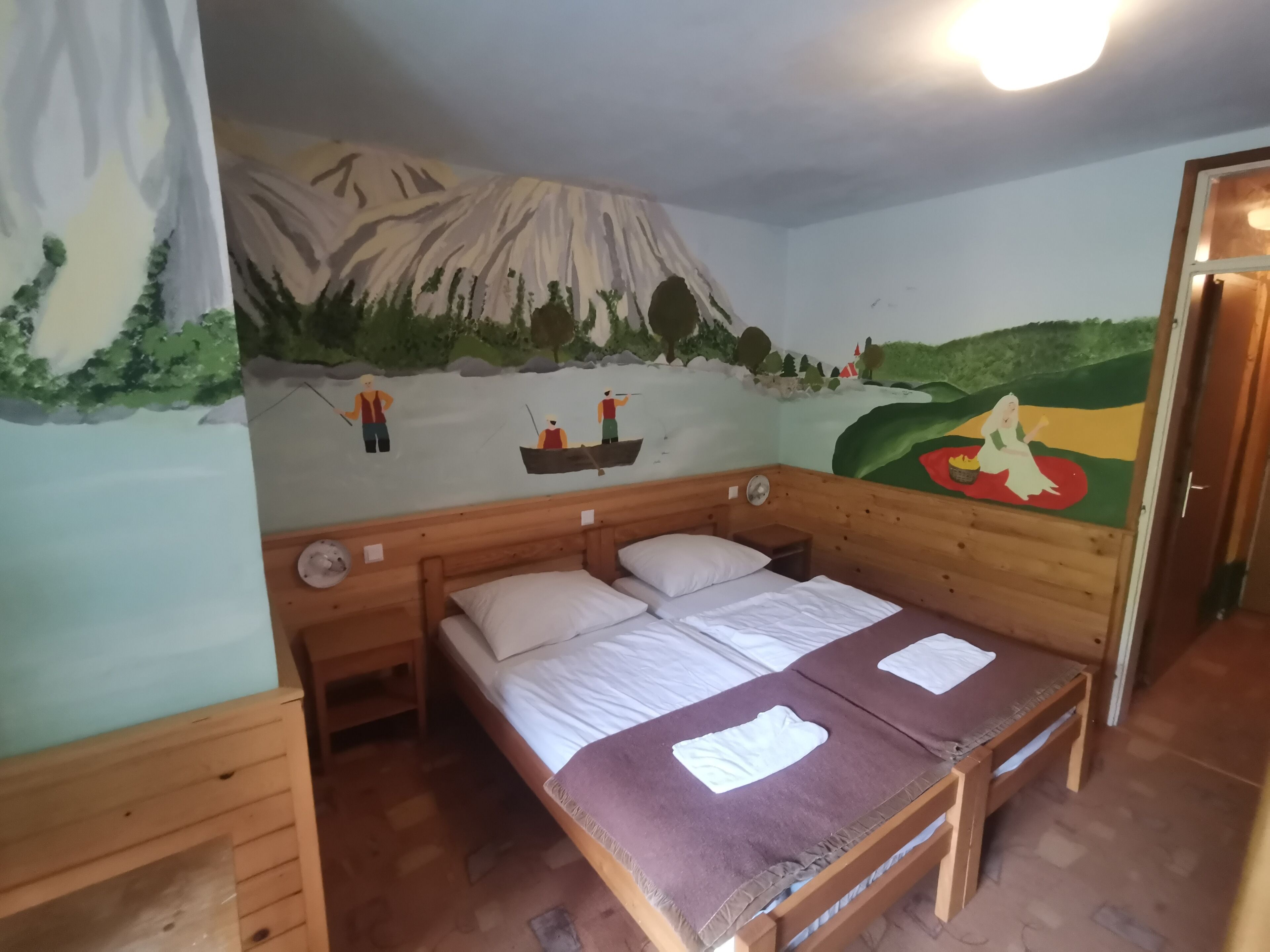 A room with a bed, a wooden headboard, a nightstand, and a wall mural depicting a mountain scene with people fishing.
