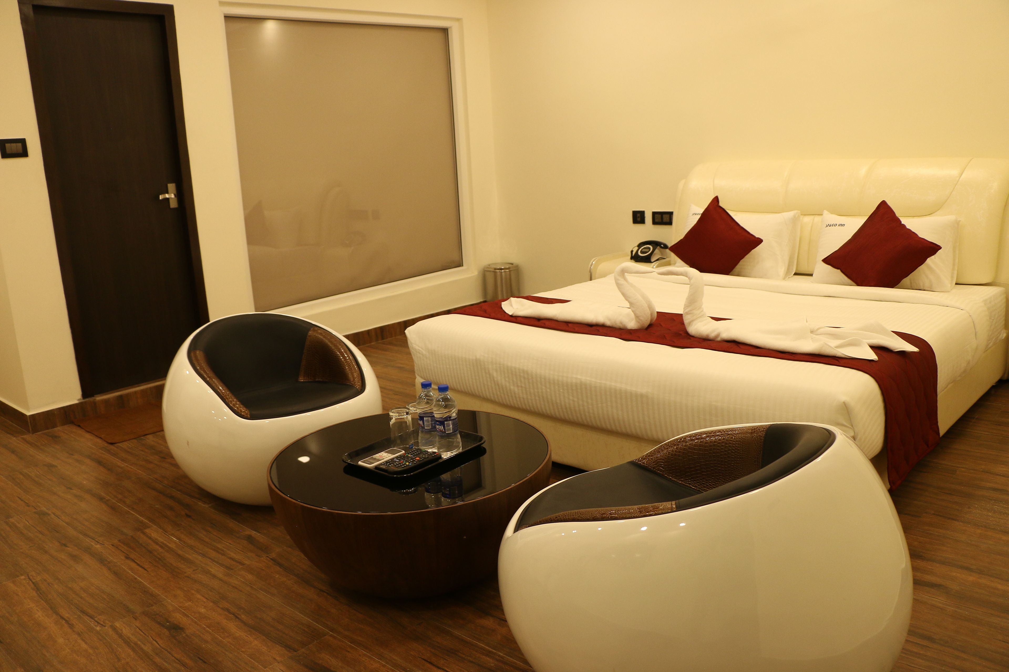 A hotel room with a bed, two white armchairs, a round coffee table with a remote control and water bottles, and a door with a light switch.