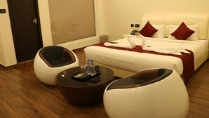 Une chambre d’hôtel avec un lit, deux fauteuils blancs, une table basse ronde avec une télécommande et des bouteilles d’eau, et une porte avec un interrupteur.