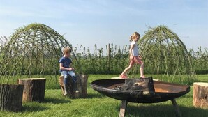 Dos niños jugando cerca de un brasero circular rodeado de troncos de madera en un campo de césped, con grandes estructuras verdes al fondo.