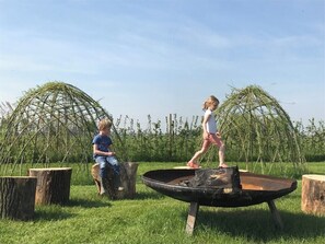 Children play near a fire pit surrounded by wooden stumps in a grassy area.