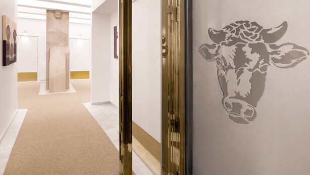 Un pasillo de diseño moderno, con una puerta de vidrio con marco dorado, alfombra color beige y una pared con silueta de vaca.