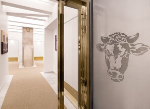 Un couloir au design moderne, avec une porte en verre dotée d’un cadre doré, un tapis beige et un mur orné de la silhouette d’une vache.