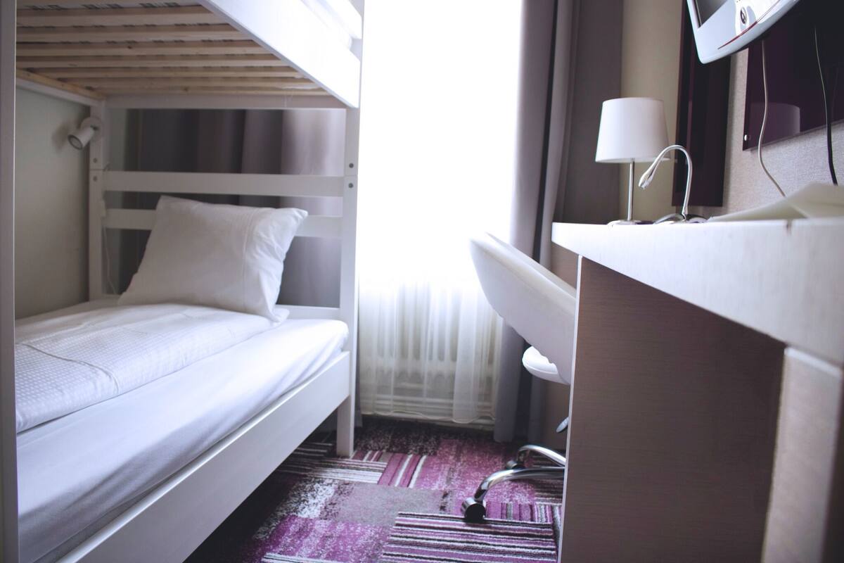 Una cama individual con un colchón y almohada blancos, un escritorio con una silla blanca, y una lámpara de escritorio pequeña.