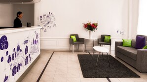 Zona de recepción de un hotel con diseño moderno, que incluye un mostrador de recepción, área de descanso con sillas y un sofá, una araña y una pared decorativa con ilustraciones.