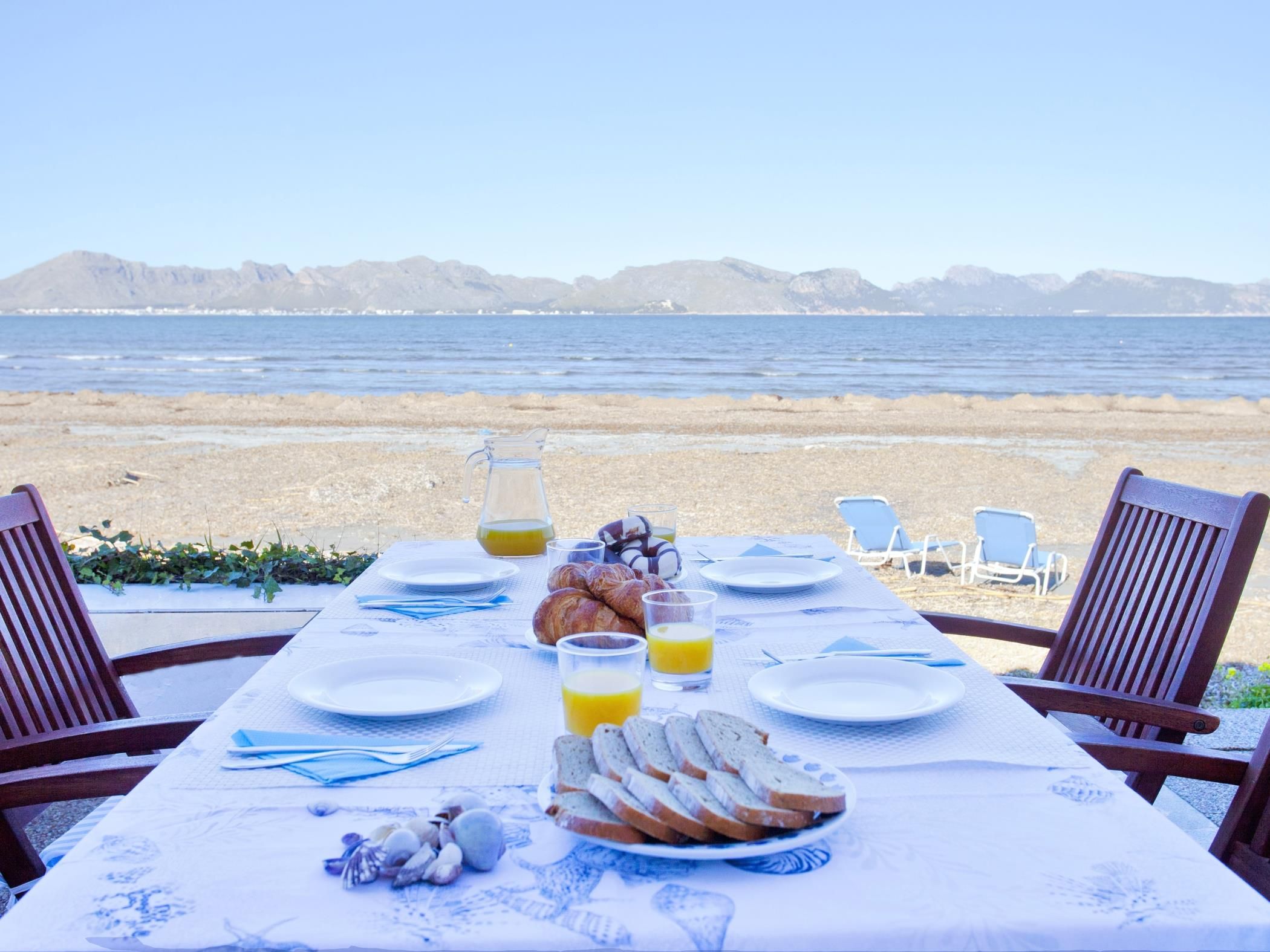 A table set for breakfast with croissants, bread, and juice glasses, overlooking a beach with mountains in the distance.