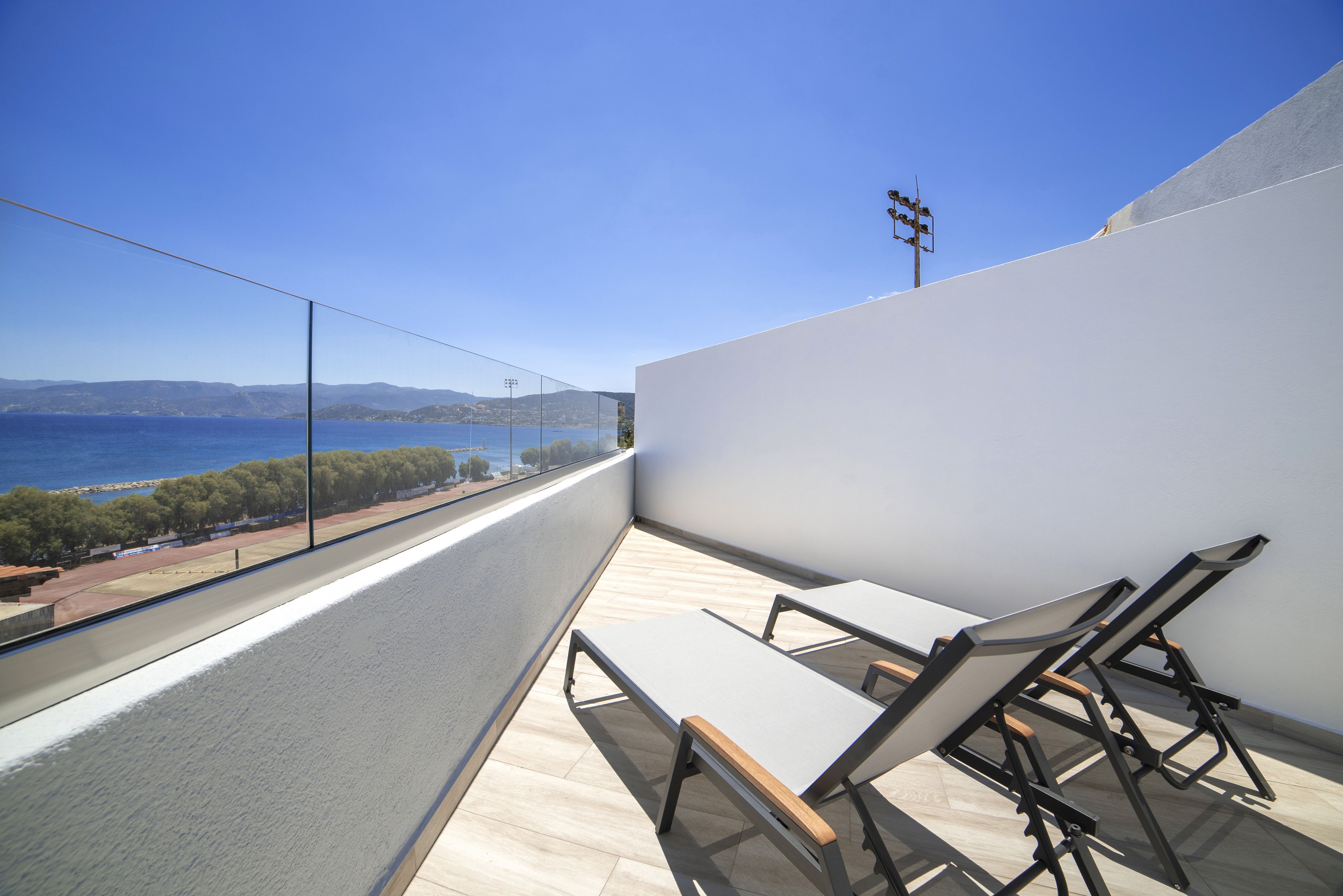 A balcony with a view of the sea and mountains, featuring two sun loungers and a glass barrier.