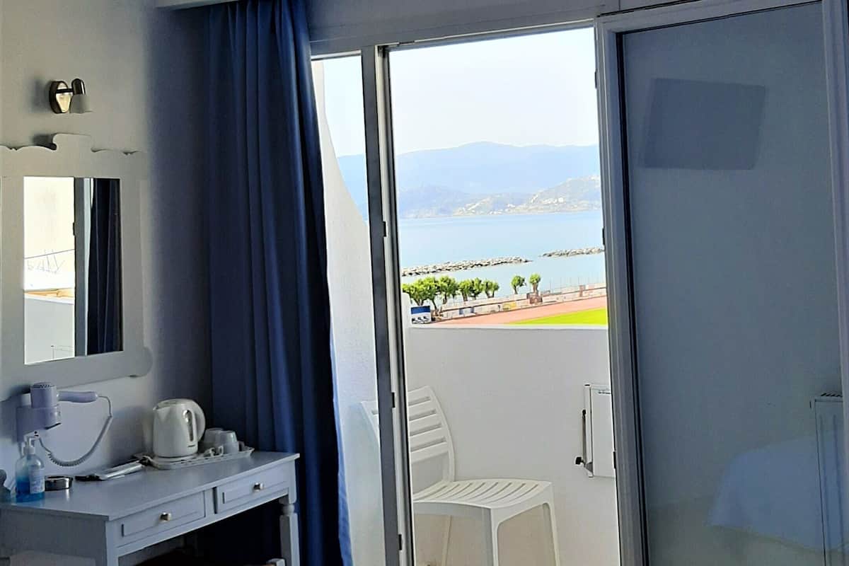 Habitación de hotel con cama, escritorio con teléfono y tetera, una silla y vista al mar a través de una puerta corrediza de vidrio.