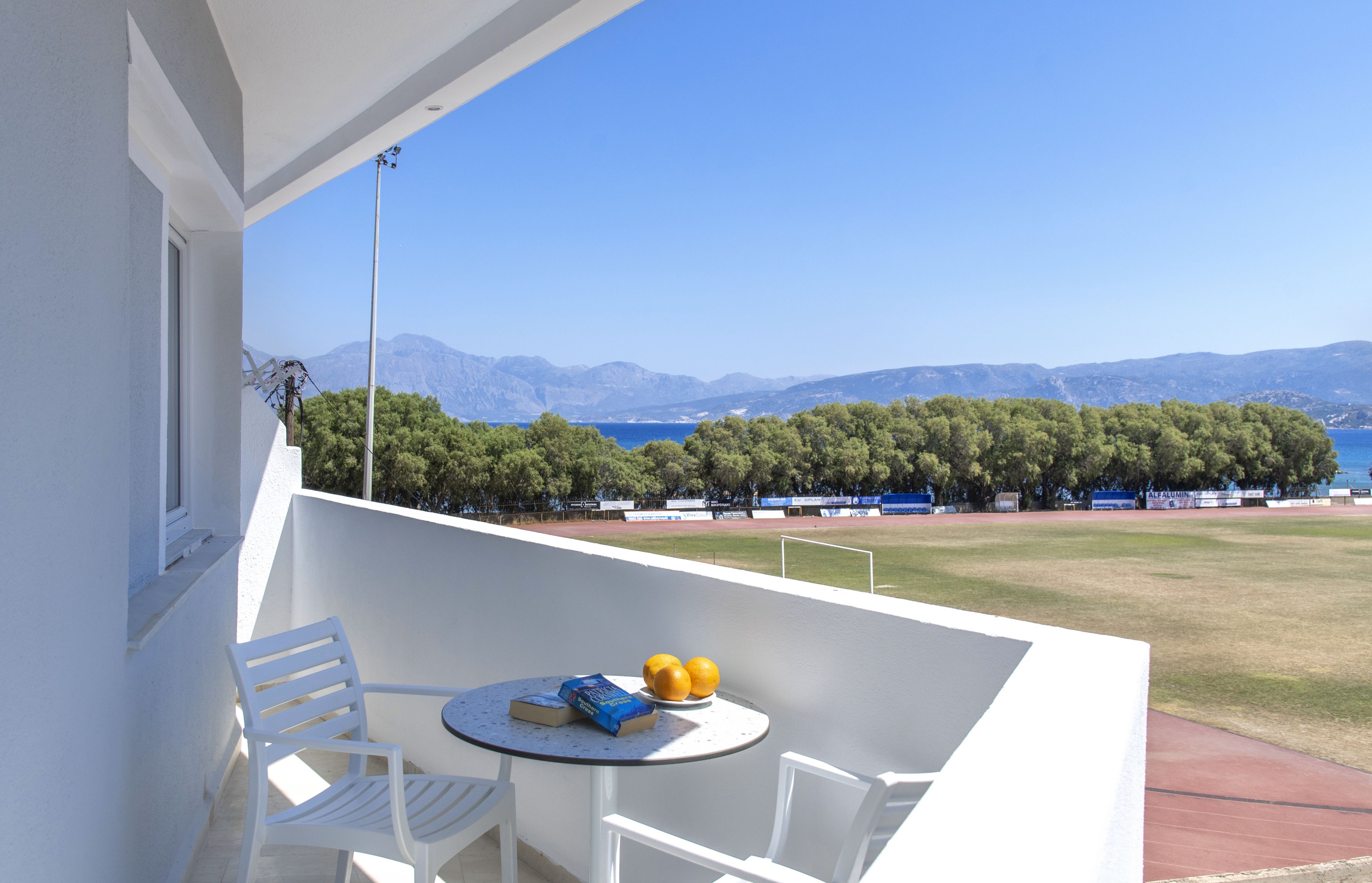 A balcony with white chairs and a small table, overlooking a sports field and a clear blue sky.