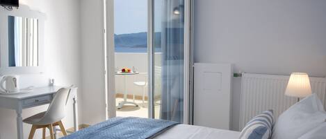 Un dormitorio con cama, un escritorio y una silla. Se ve el exterior a través de un ventanal.
