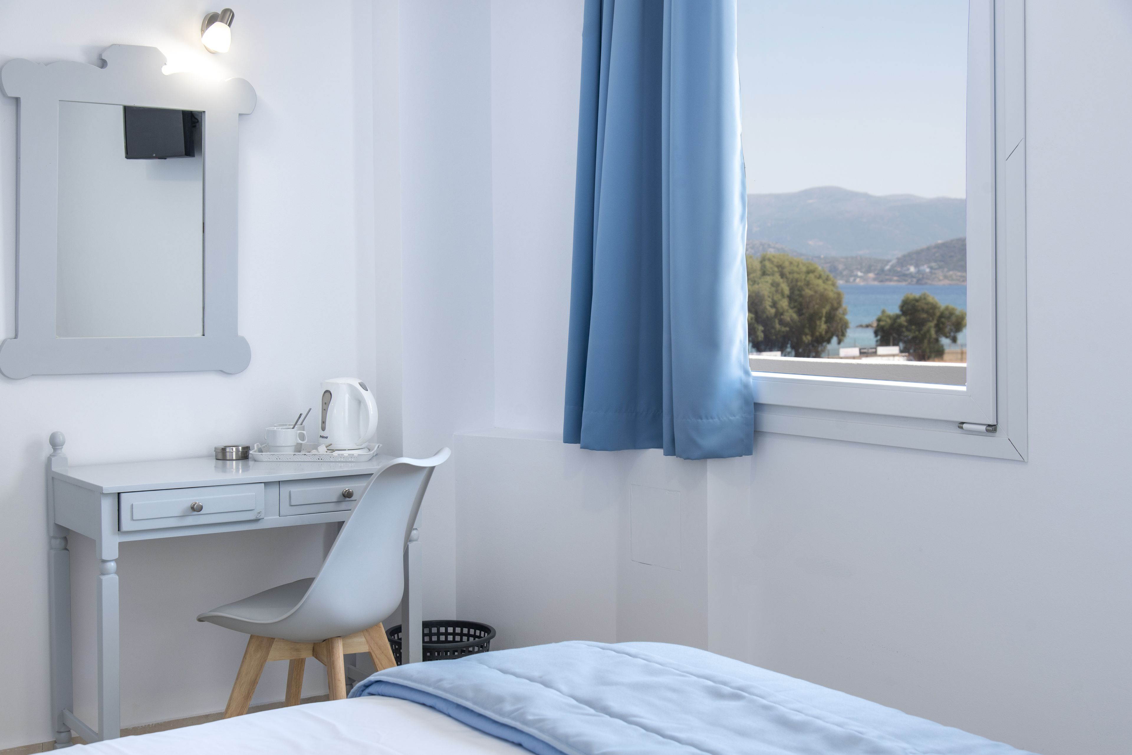A bedroom with a bed, a desk with a chair, a window with a view of the sea and hills, and a wall-mounted TV.