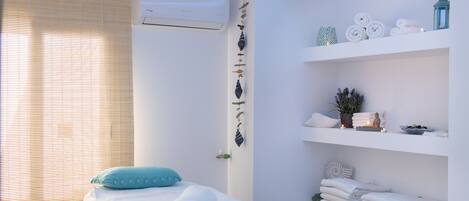 A spa room with a bed, towels, a small table with a cup and a brush, and shelves with towels and plants.