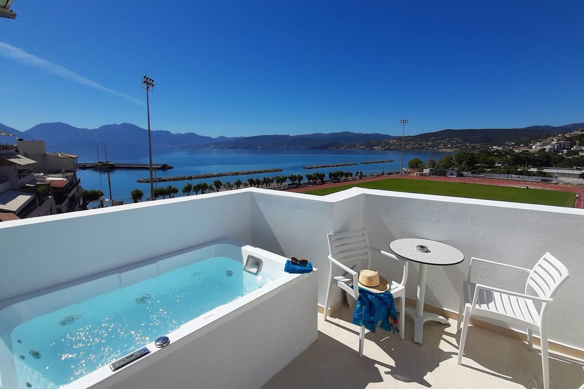 Un balcón con piscina, una mesita y una silla, con vistas a la costa, montañas y un puerto deportivo.