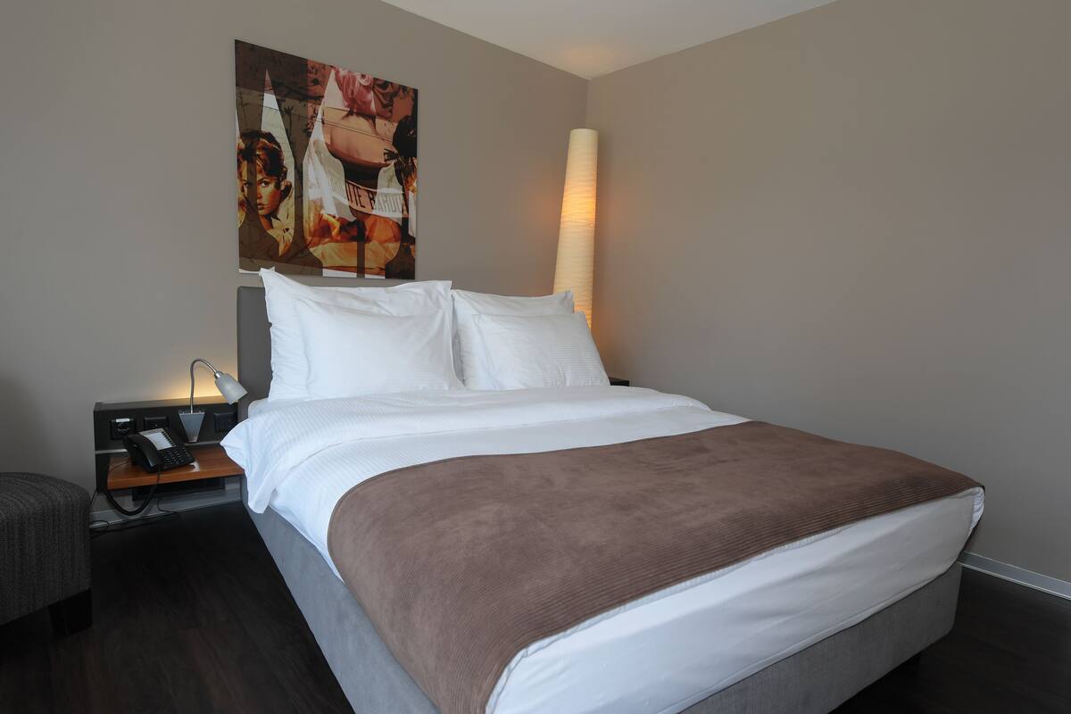 Una cama bien hecha con una manta marrón, una lámpara de noche y un teléfono sobre una mesita de madera.
