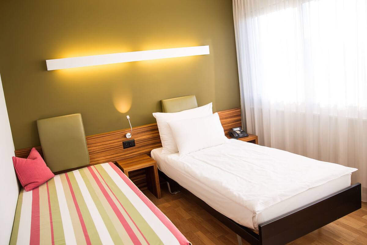 Habitación de hotel con cama, mesitas de noche, lámpara, teléfono y ventana con cortinas.