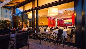 Un espace extérieur aménagé pour les repas, avec des tables et des chaises, de grandes fenêtres et un éclairage intérieur visible à travers le verre.
