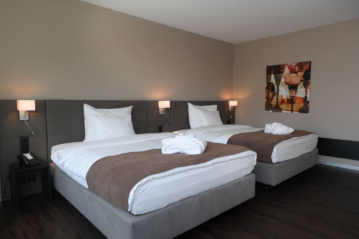Una habitación de hotel con dos camas, cada una con ropa blanca y mantas marrones, una mesita de noche con teléfono y lámparas fijadas a la pared.