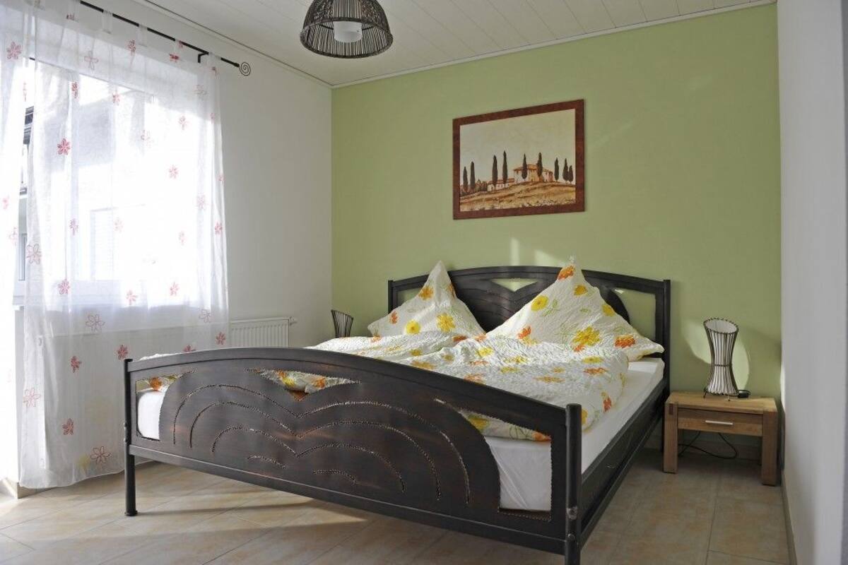 Um quarto com cama, mesa de cabeceira, luminária, janela com cortinas e um quadro na parede.
