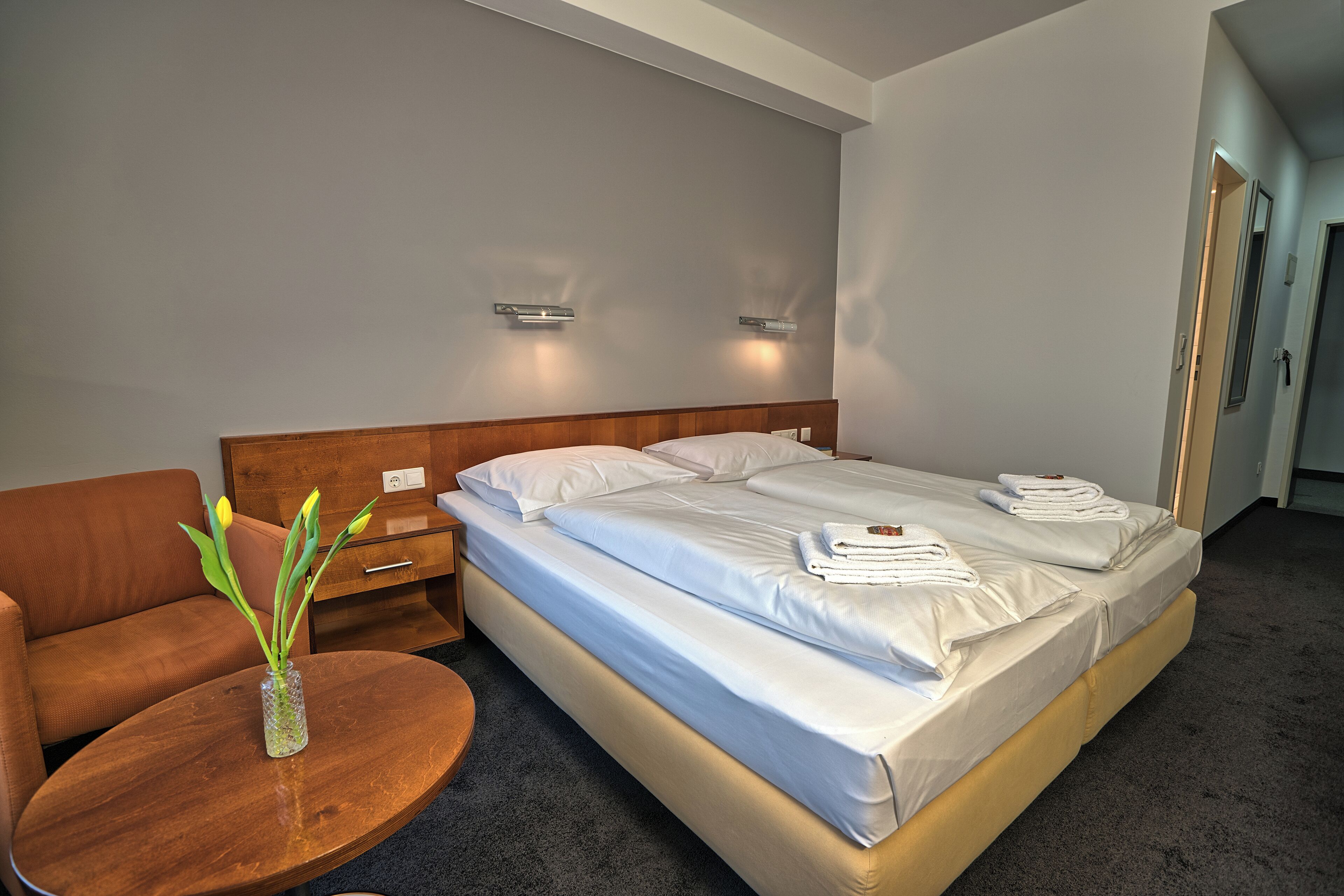 A hotel room with a bed, a wooden nightstand, a round table with a vase of flowers, and a brown sofa.
