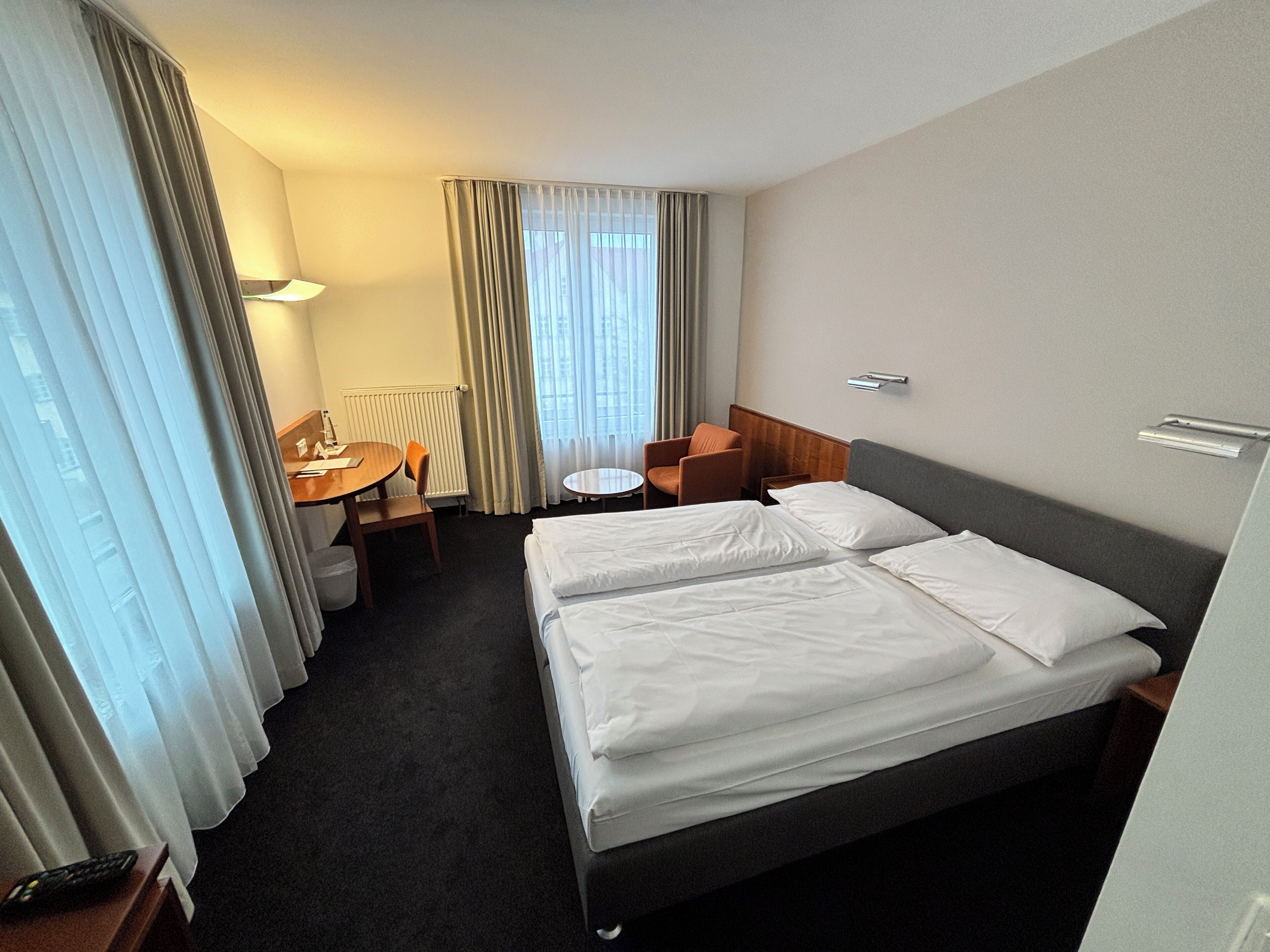 A hotel room with two beds, a desk, a chair, a small table, and a window with curtains.