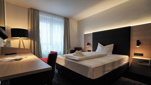 Habitación de hotel moderna con una cama bien hecha, un escritorio con lámpara y vistas al exterior a través de grandes ventanales.