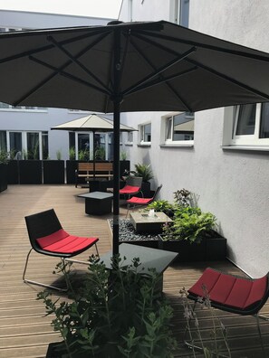 An outdoor patio area with a wooden deck, black and red lounge chairs, a table with a fountain, and potted plants. - Hotel4you (Dorfen)