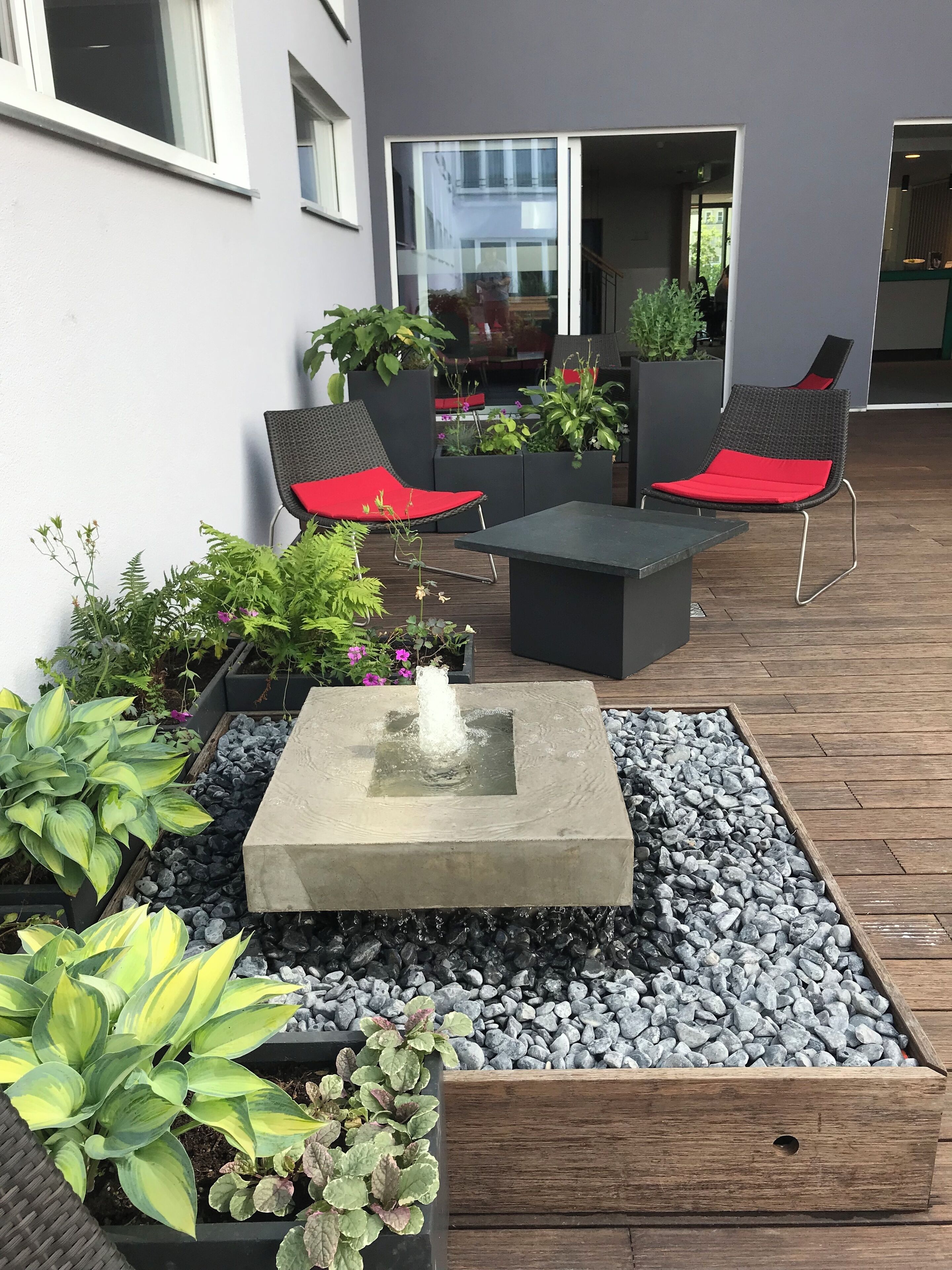 Un patio al aire libre con una mesa de piedra, dos sillas de mimbre con cojines rojos y varias macetas con plantas.