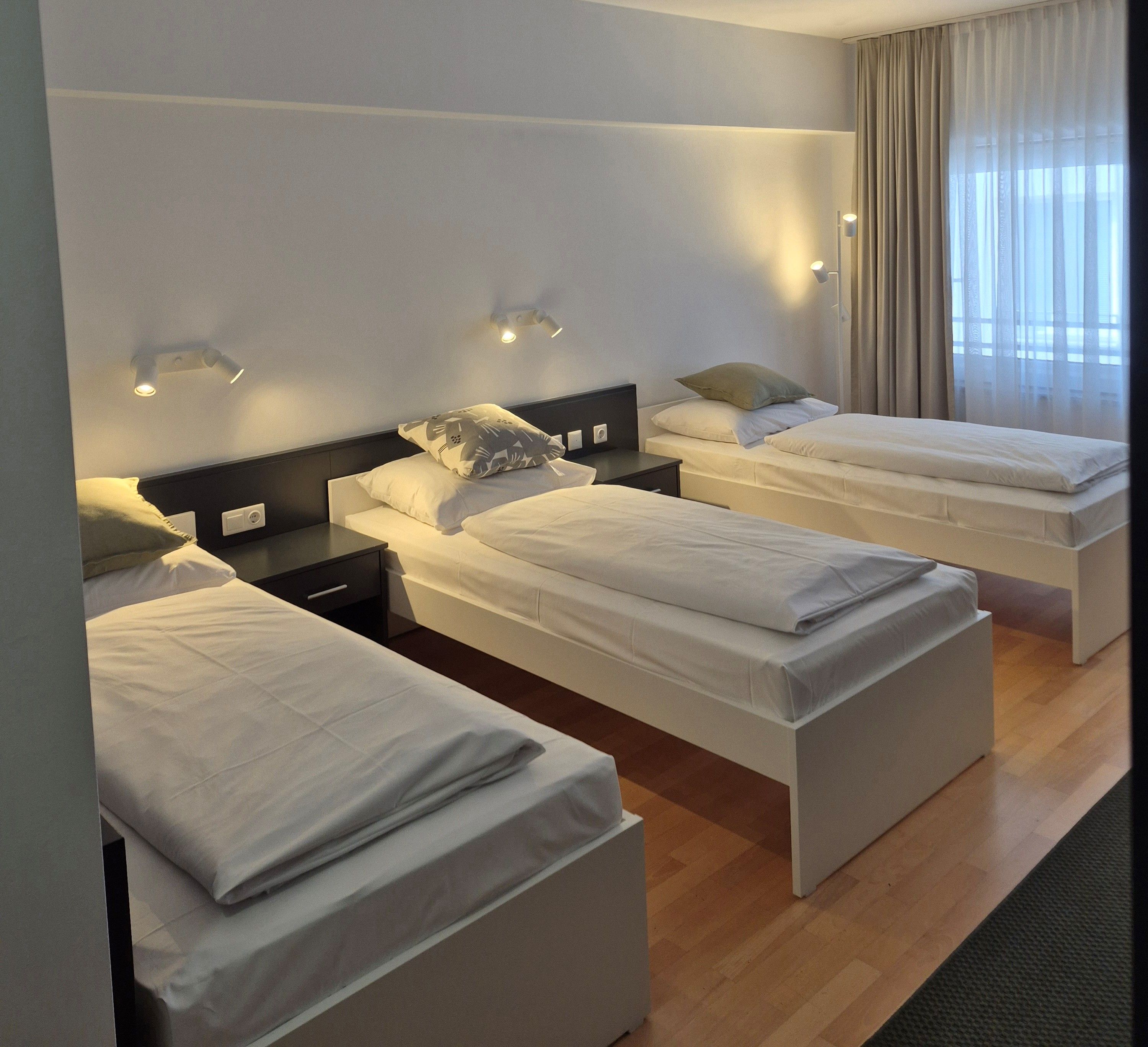 A hotel room with three single beds, a nightstand, a wall-mounted lamp, and a window with curtains.