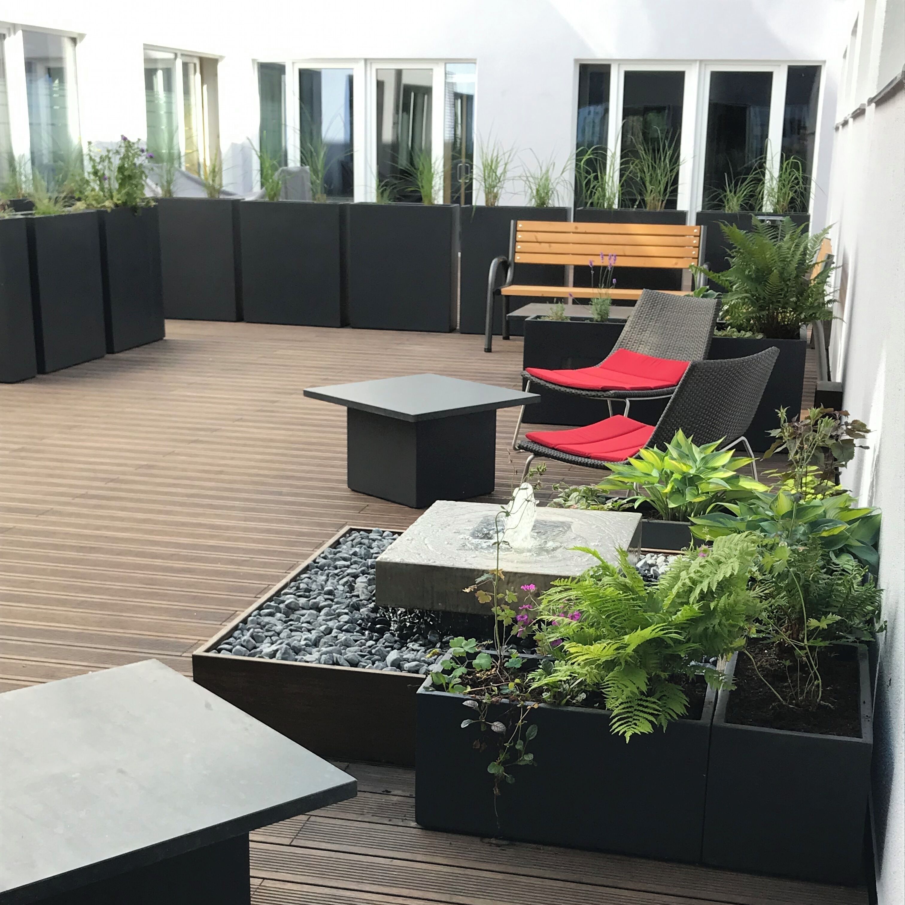 En tagterrasse med træterrasse, et sort bord, en bænk med røde hynder og forskellige plantekasser.