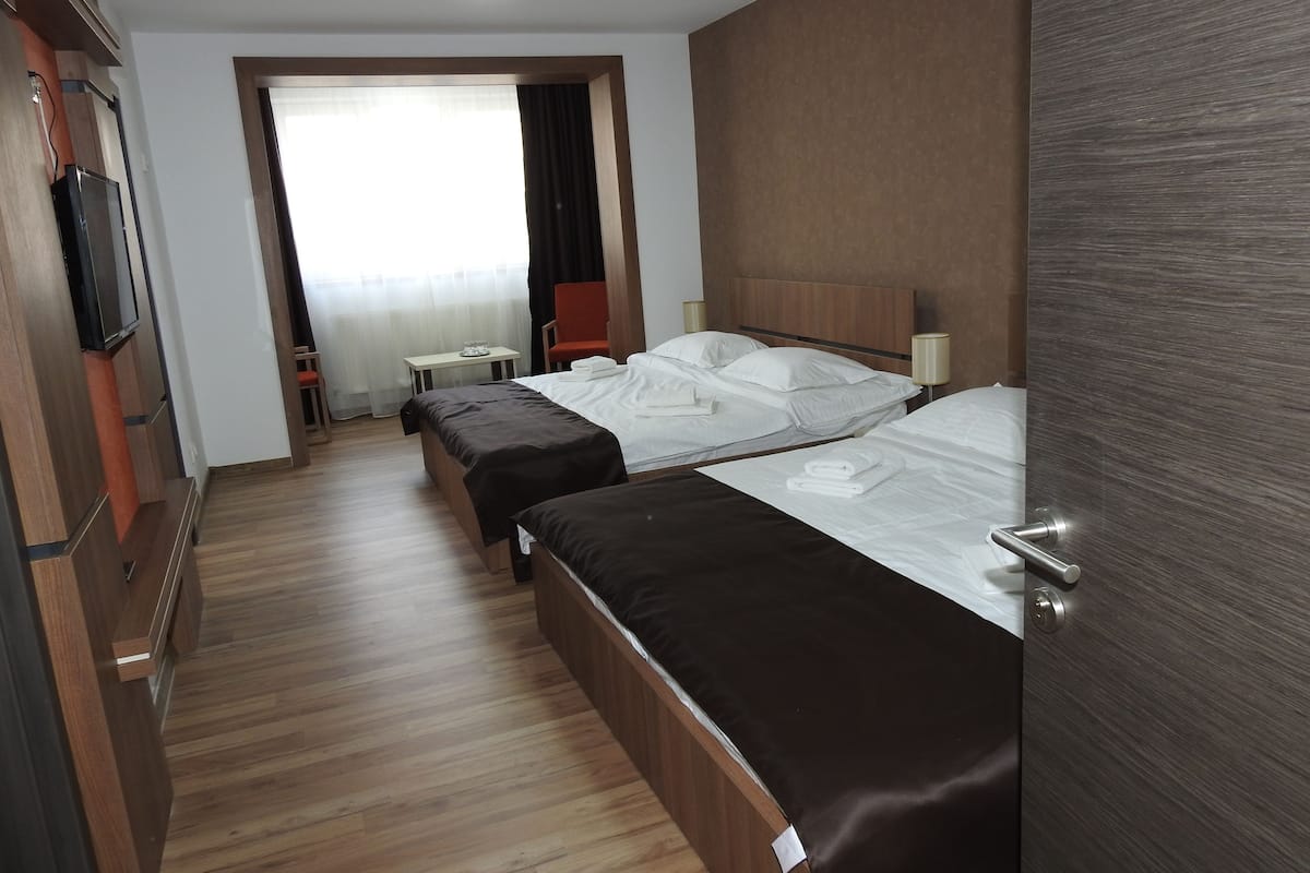 Habitación de hotel con dos camas, piso de madera, televisión, una mesita y ventana con cortinas.