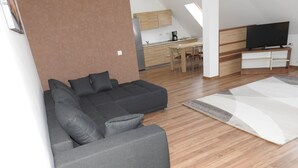 Una sala de estar moderna con un sofá seccional gris, un televisor de pantalla plana sobre un soporte de madera, y una cocina al fondo.