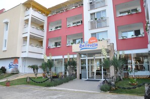 A hotel named 'Belleville' with a red and white facade, multiple balconies with potted plants, and a well-maintained entrance area with a chain-link fence and landscaped garden. - Hotel BelleVille (Sunny Beach)